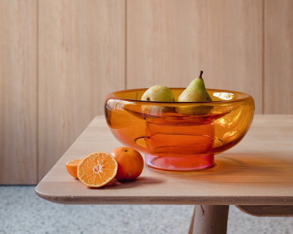 semi-transparent orange cylinder glass base holding a semi-transparent deep yellow moon shape glass forming a bowl on a wooden table with fruits around