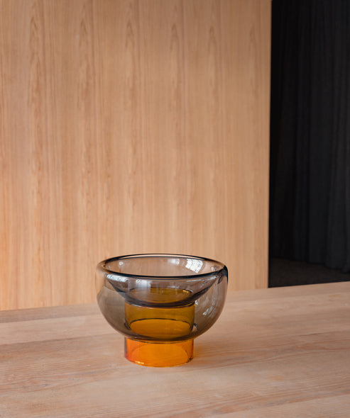 semi-transparent yellow cylinder glass base holding a semi-transparent grey moon shape glass forming a bowl on a cement floor in front of a wooden wall
