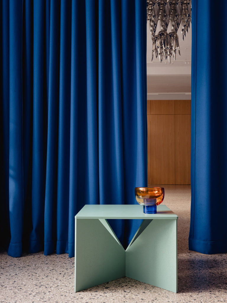 semi-transparent deep blue cylinder glass base holding a semi-transparent orange moon shape glass forming a bowl on a mint table terrazzo floor and blue curtains behind